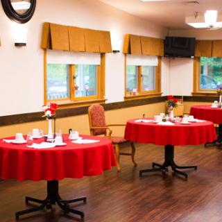 A cozy dining area featuring tables dressed in red tablecloths, set for a meal with decorative touches and natural light coming through the windows.