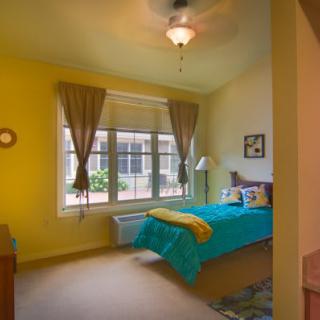 A cozy bedroom with yellow walls and a turquoise bedspread, featuring large windows with curtains and a ceiling fan.