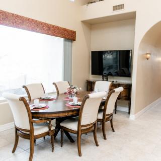 A well-lit dining area with a wooden table and chairs, featuring a large window and a television mounted on the wall.