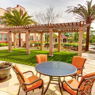 A sunny patio area with a table and orange chairs, surrounded by a pergola and palm trees.