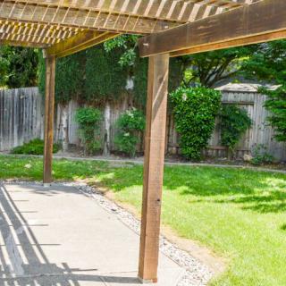 A backyard with a wooden pergola casting shadows on the patio, surrounded by a green lawn and a wooden fence.