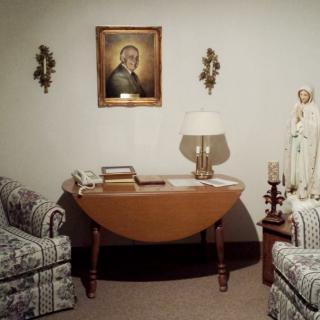 A cozy living room with a central wooden table, flanked by two armchairs. A portrait hangs on the wall above the table, accompanied by decorative elements such as a lamp and a statue.