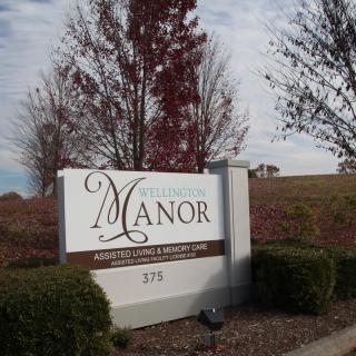 A sign for a place named 'Manor' surrounded by trees and bushes.