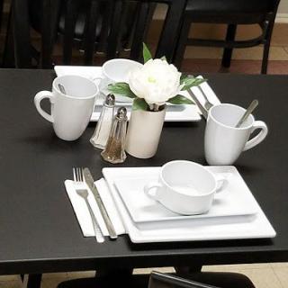 A neatly set dining table with white dishes, mugs, silverware, and a floral centerpiece.