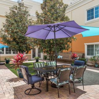 A cozy outdoor patio area with a purple umbrella, chairs, and a table surrounded by green plants.