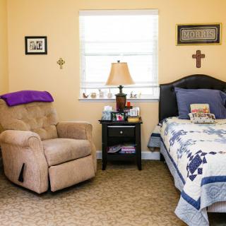 A cozy bedroom featuring a comfy armchair, a neatly made bed with a decorative quilt, and a nightstand with personal items.