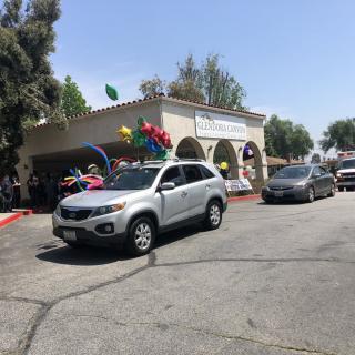 A parade of cars decorated with balloons passes by a building on a sunny day.