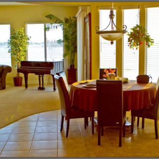 A spacious living and dining area with a grand piano, surrounded by large windows and indoor plants.