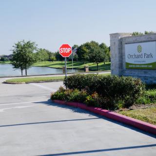 Entrance to a residential community with a stop sign, landscaped greenery, and a view of the lake.