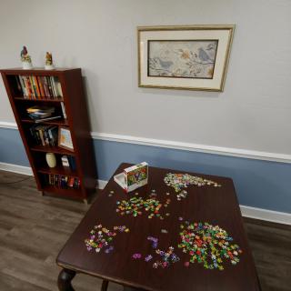 A room with a partially completed puzzle on a table, a bookshelf with books and decorations, and a painting on the wall.