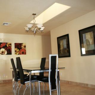 A modern dining room with a glass table and black chairs, adorned with a chandelier and framed artwork on beige walls.