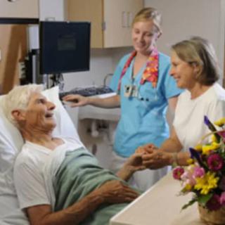 A nurse stands by a patient in a hospital bed while a visitor holds the patient's hand.