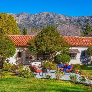 A charming house with a red-tiled roof is set against a backdrop of mountains, surrounded by lush gardens and trees, with colorful patio furniture in the foreground.