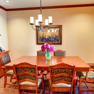 A cozy dining room with a wooden table, surrounded by chairs, a floral centerpiece, and an elegant chandelier overhead.