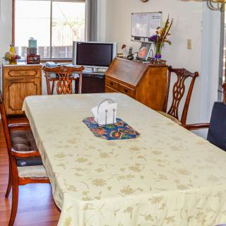 A cozy dining room with a table covered by a tablecloth, surrounded by wooden chairs and featuring a computer setup near the window.