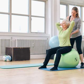 An elderly person exercising on a yoga ball with guidance from a personal trainer in a bright indoor studio.