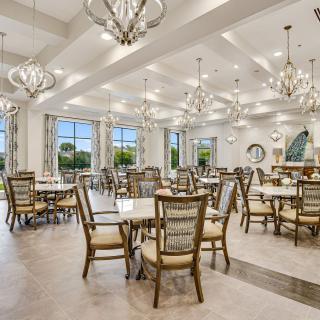 A spacious and elegantly designed dining room with chandeliers, featuring multiple tables and chairs under bright lighting.