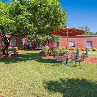 A serene outdoor garden space featuring a seating area under an umbrella, surrounded by lush greenery and a charming brick building.