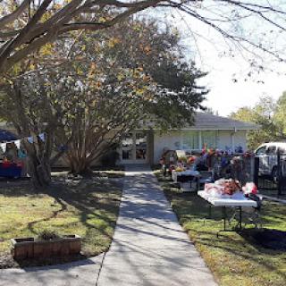 A bustling yard sale outside a house, with various items displayed along the pathway under the shade of trees.