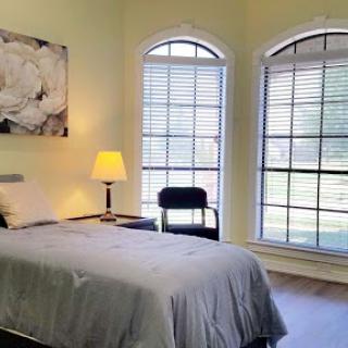 A cozy bedroom featuring a single bed, a lamp, and two large windows that allow natural light to fill the space.