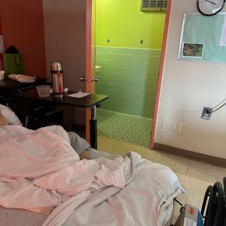 A view of a hospital patient room featuring a bed and a nearby wheelchair, with a brightly colored bathroom in the background.