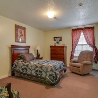 A cozy bedroom with a bed, dresser, and armchair near a window with red curtains.