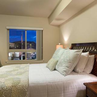 A cozy bedroom showcasing a neatly made bed with decorative pillows, illuminated by soft lighting and a window revealing the evening sky.