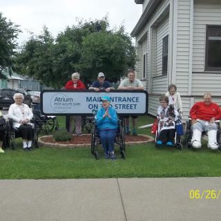A group of elderly individuals sitting outside near a building with a sign, some in wheelchairs.