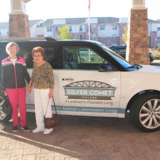 Two people standing in front of a car with a 'Silver Connect' logo, outside a building.