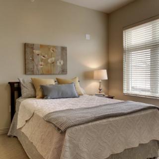 A cozy bedroom with a neatly made bed, side lamp, and large window providing natural light.
