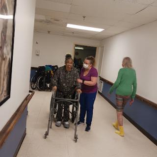 A caregiver assists a patient in a wheelchair while another caregiver walks nearby in a hospital hallway.