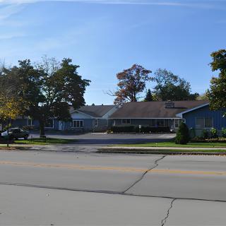 A one-story building with a parking lot, surrounded by trees, viewed from across the street on a clear day.