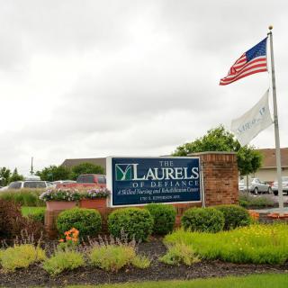 The entrance sign of The Laurels of Defiance nursing home with an American flag waving under a cloudy sky.
