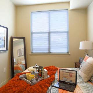 A cozy bedroom with a neatly arranged bed, featuring a laptop, mirror, and window.