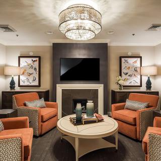 A modern living room with orange chairs, a central coffee table, and a chandelier above, features a fireplace and television.