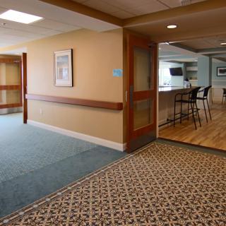 A hallway with carpeted flooring leading to a kitchen area with bar stools.