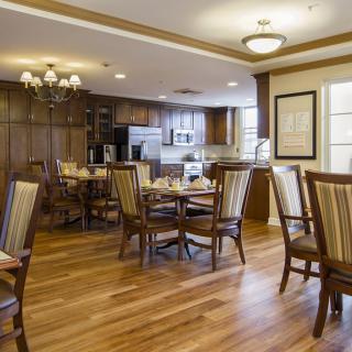 A cozy dining room with wooden flooring, neatly arranged tables and chairs, and a kitchen area in the background.