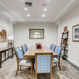 A modern dining room with a wooden table, blue upholstered chairs, artwork on the walls, and a bookshelf in the corner.