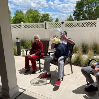 A group of people relaxing on a sunny patio, seated on chairs near a white fence.