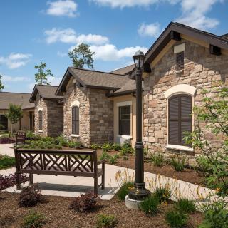 A charming residential building with a stone facade, surrounded by landscaped gardens, featuring a pathway leading past a decorative bench and lamp post on a sunny day.