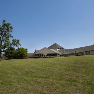 A large building with a sloping roof surrounded by an expansive lawn and trees under a clear blue sky.