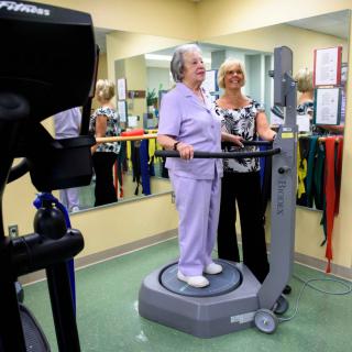 An elderly woman is being assisted by a healthcare worker while using an exercise machine in a physical therapy setting.