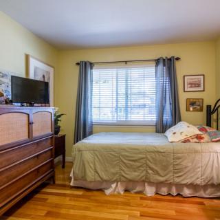 A cozy bedroom with a bed, wooden dresser, and a television next to a window with curtains.