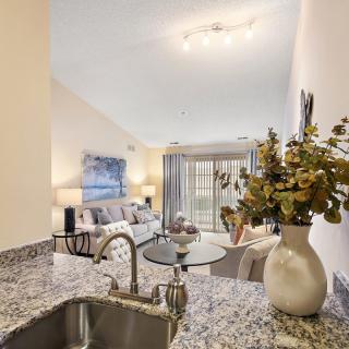 A modern open-plan living area featuring a stylish kitchen with granite countertops and a cozy living room adorned with neutral tones and decorative flowers.