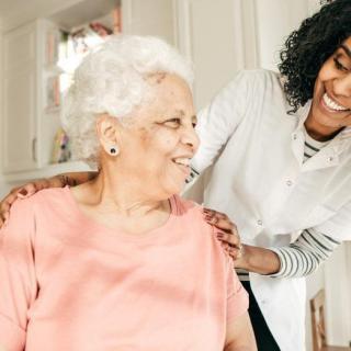 A caregiver is smiling and supporting an elderly woman at home.