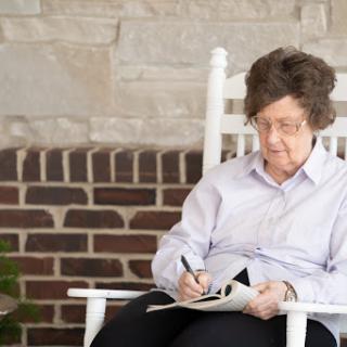An elderly woman sitting in a rocking chair, focused on writing something down.