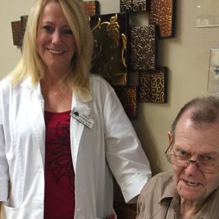 A nurse is standing next to an elderly man in a medical setting, both smiling.