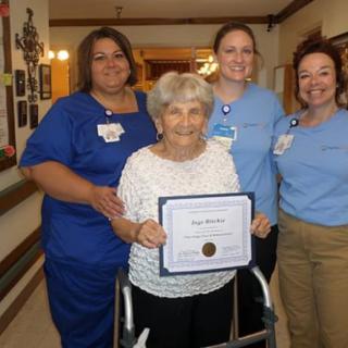 An elderly woman stands with a certificate, surrounded by nursing staff in a healthcare facility.