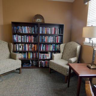 A cozy reading corner with a bookshelf, two chairs, a lamp, and a table.