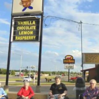A group of people sitting outside near a sign displaying ice cream flavors: vanilla, chocolate, lemon, and raspberry.